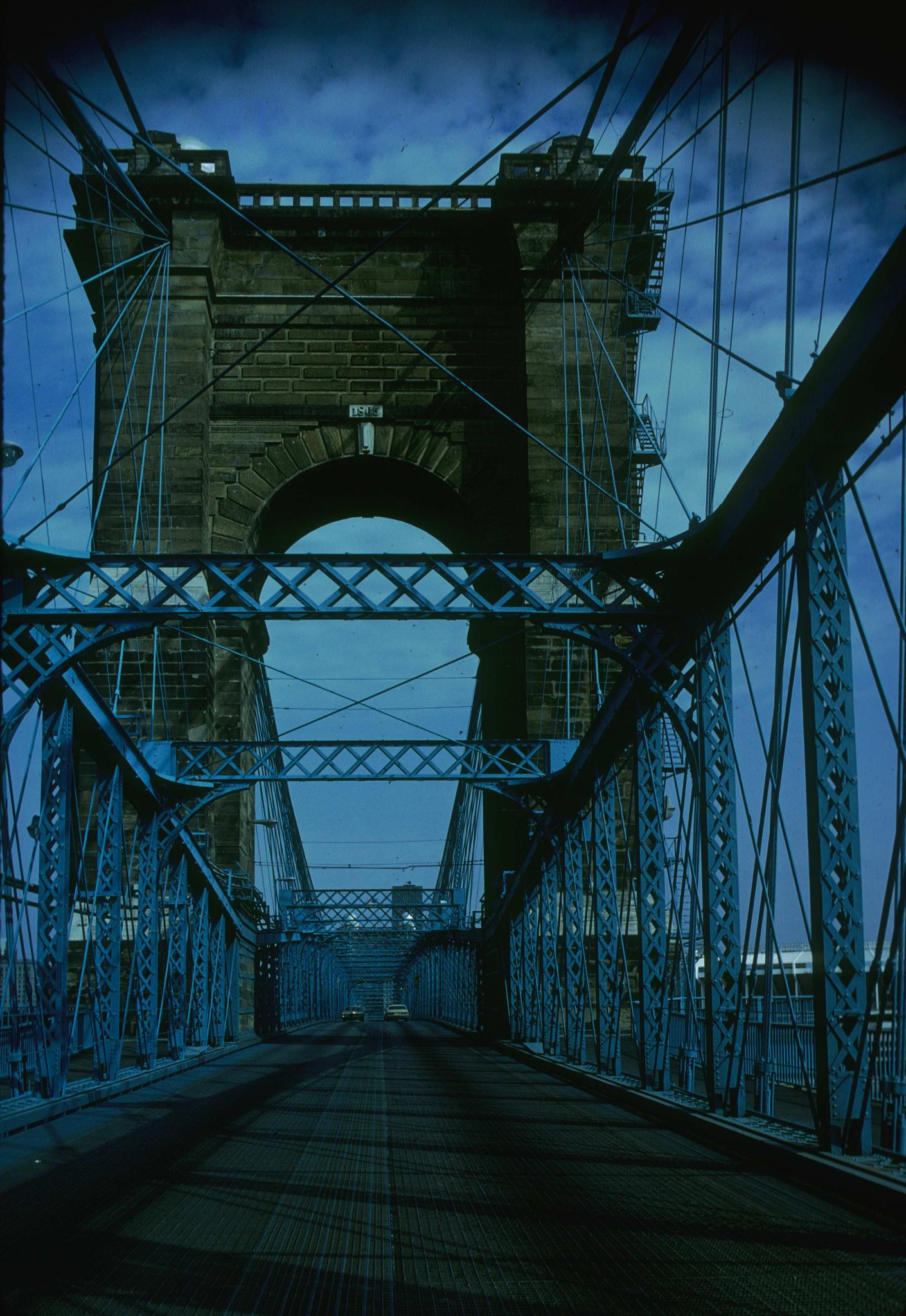 Photograph looking north along the deck of the Cincinnati Bridge.  Two cars are…