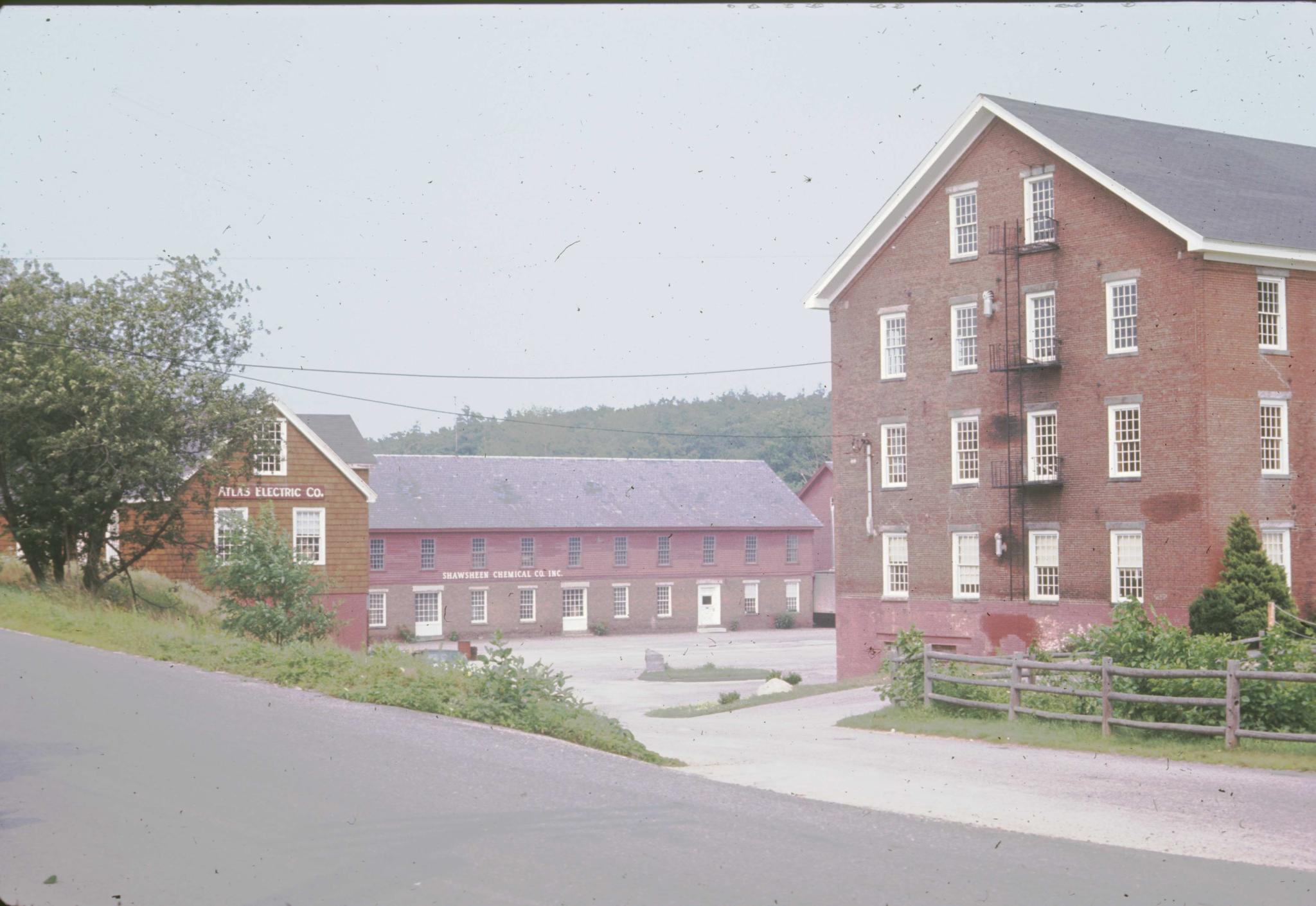 Photograph of the mill.  Business signs indicate that two of the buildings are…