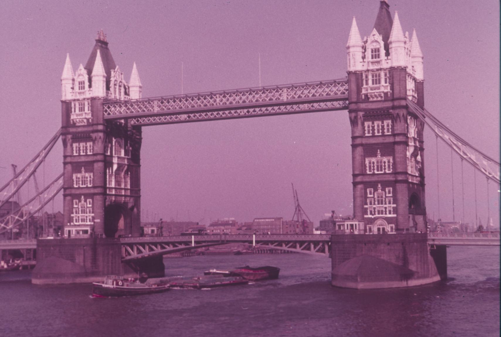 General view of Tower Bridge closed, with tugboat and barges passing beneath.