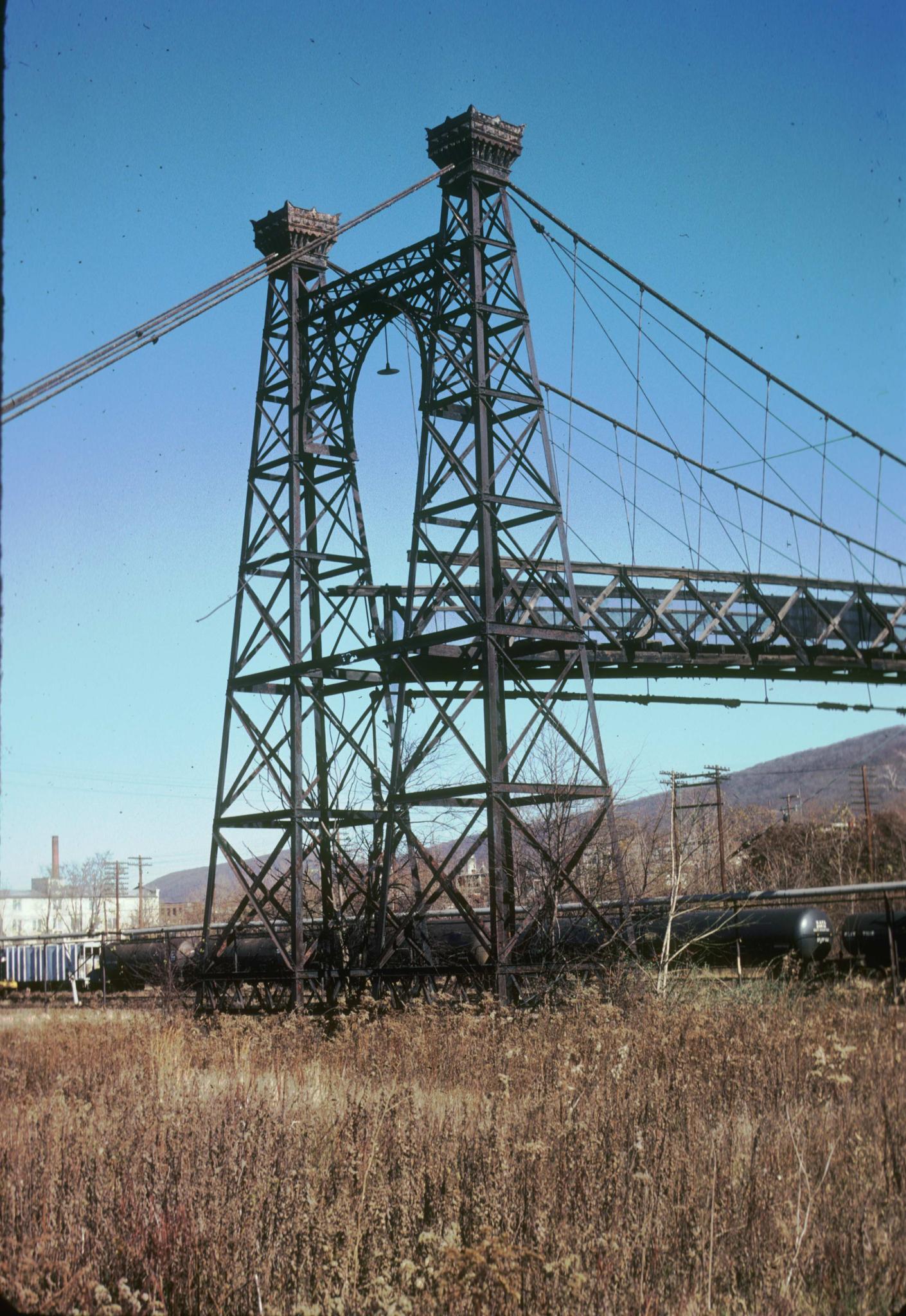 Photograph of a tower of the bridge.  A line of tanker cars is in the…