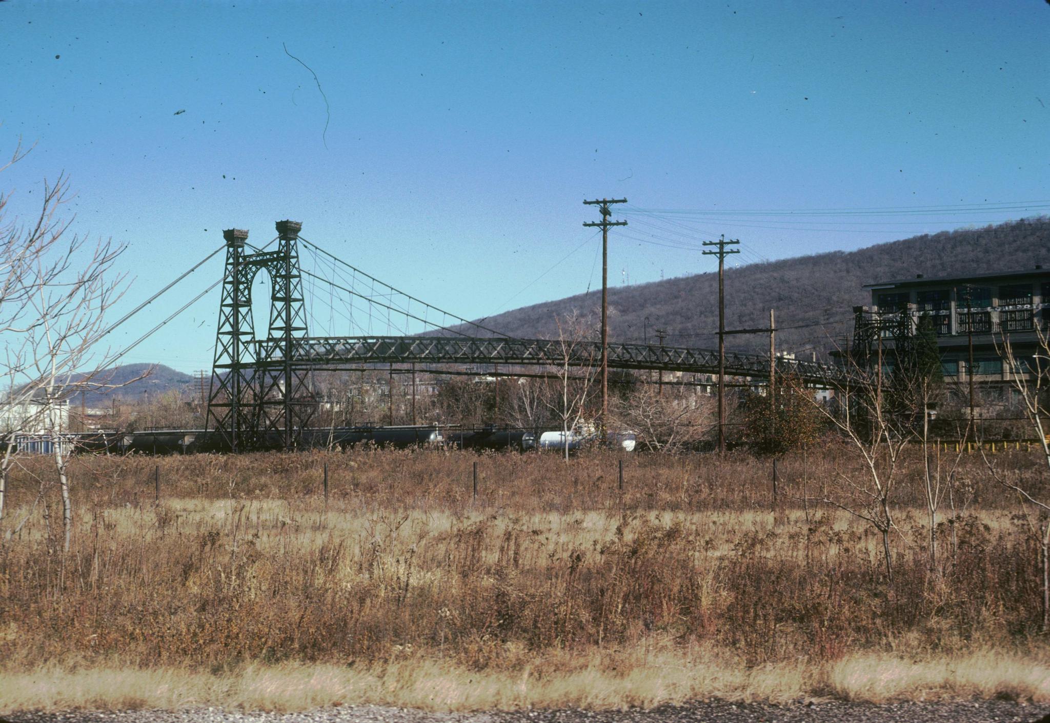 Photograph of the bridge with a line of tanker cars underneath it.